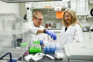 Two researchers in lab coats and goggles smile while working with vials at a lab table.