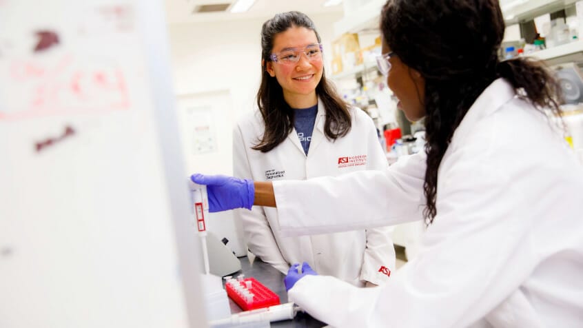 two women in Biodesign lab coats look at each other in a lab